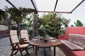 An outdoor patio with a wooden table, two chairs, a red sofa, and a variety of plants and trees in the background.