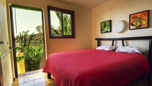 Un dormitorio con una colcha roja, una ventana con vistas a zonas verdes y dos cuadros en la pared.