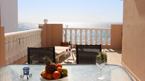 A balcony with a table set for two, featuring a fruit bowl, a wine glass, and a view of the sea.