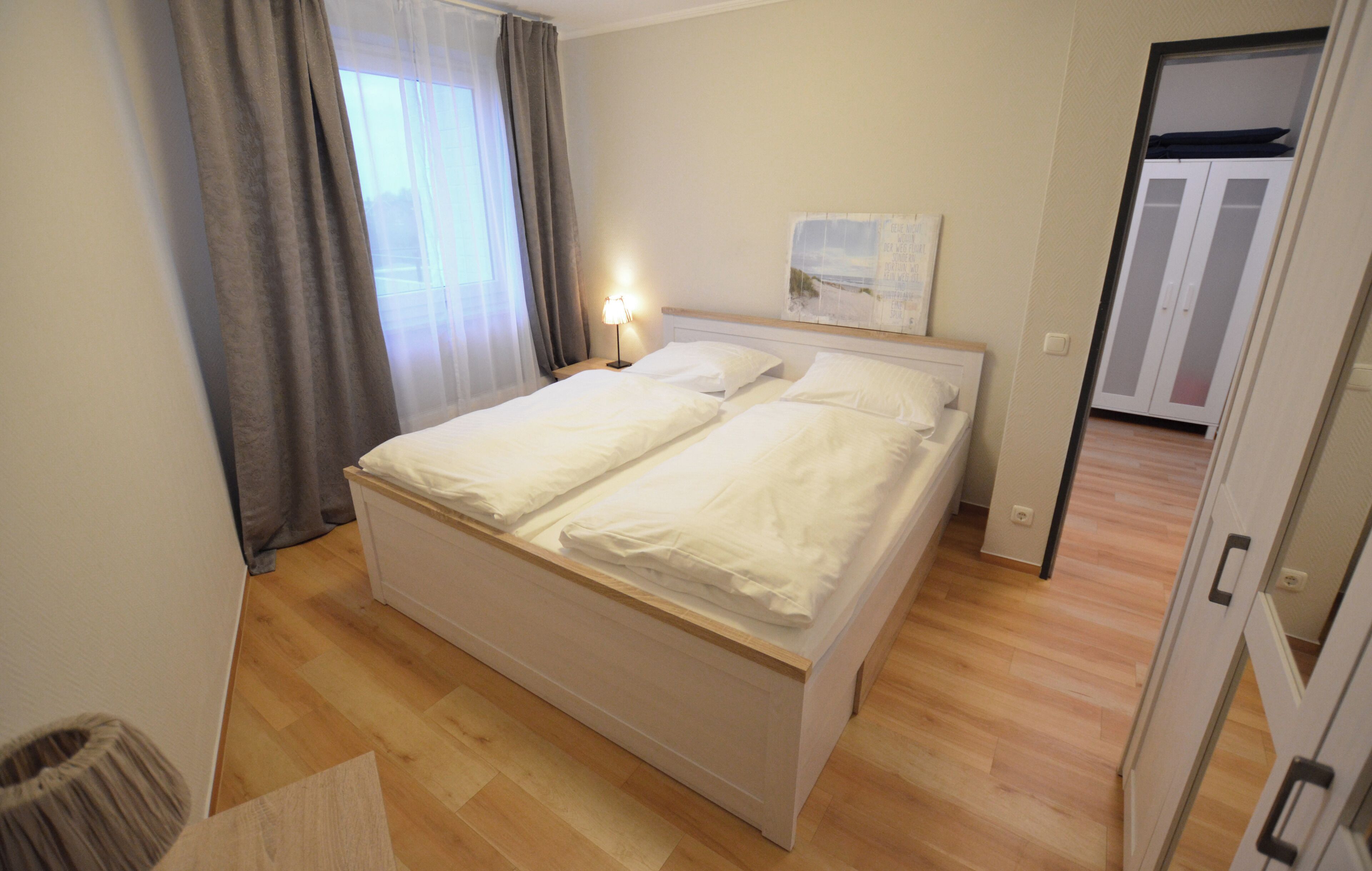 A neatly made bed with white linens in a room with wooden flooring and a window with curtains.