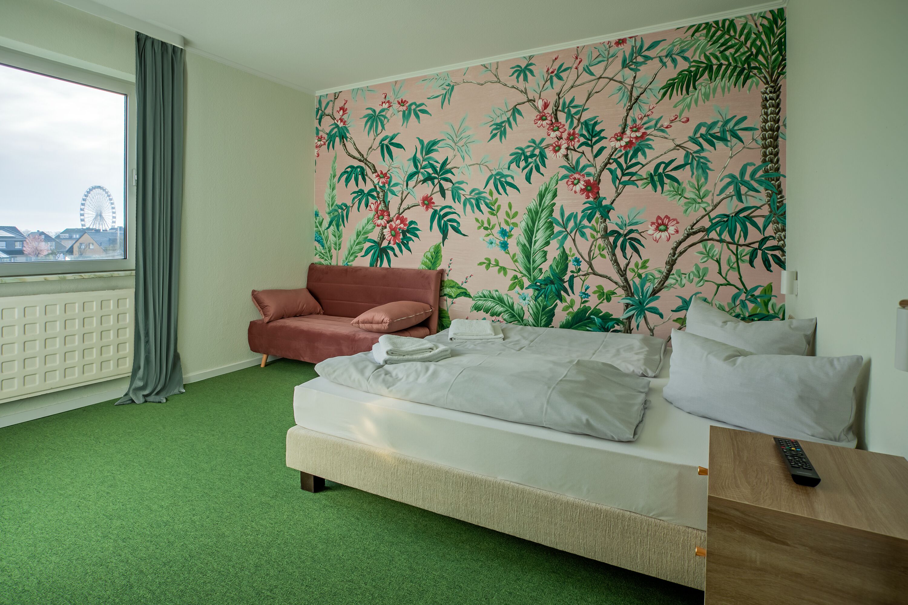 Ett hotellrum med en säng, en röd fåtölj, ett träbord med en fjärrkontroll, ett fönster med utsikt över ett pariserhjul och en blommig tapet.