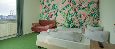 Ett hotellrum med en säng, en röd fåtölj, ett träbord med en fjärrkontroll, ett fönster med utsikt över ett pariserhjul och en blommig tapet.