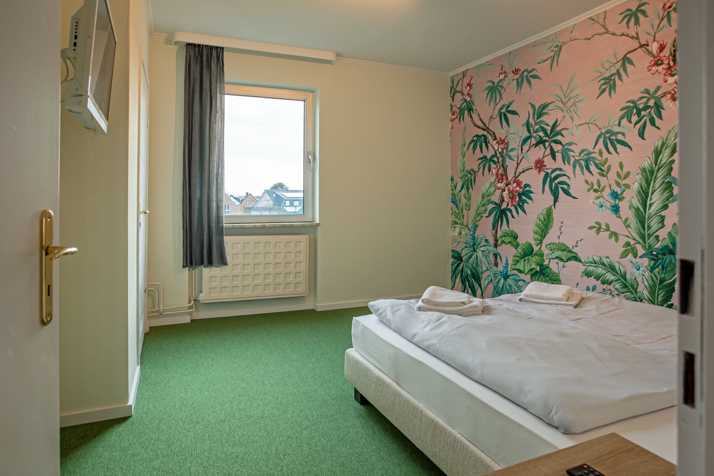 Ein Hotelzimmer mit einem Bett, einem Fenster mit Vorhängen, einem Heizkörper und floralem Wandpapier.