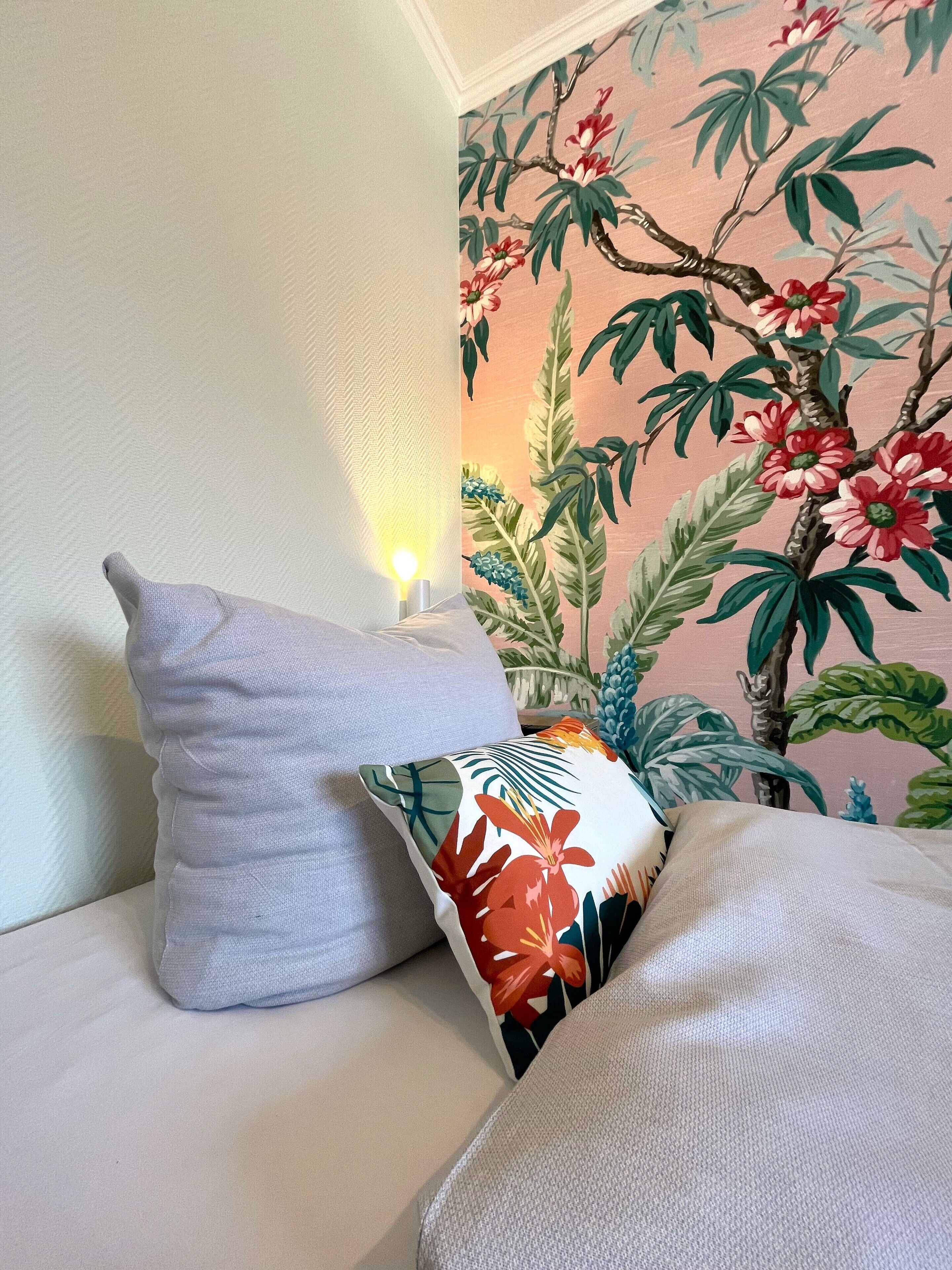 Un sofá con dos almohadas, una lisa y otra con estampado floral, frente a una pared con un mural tropical.