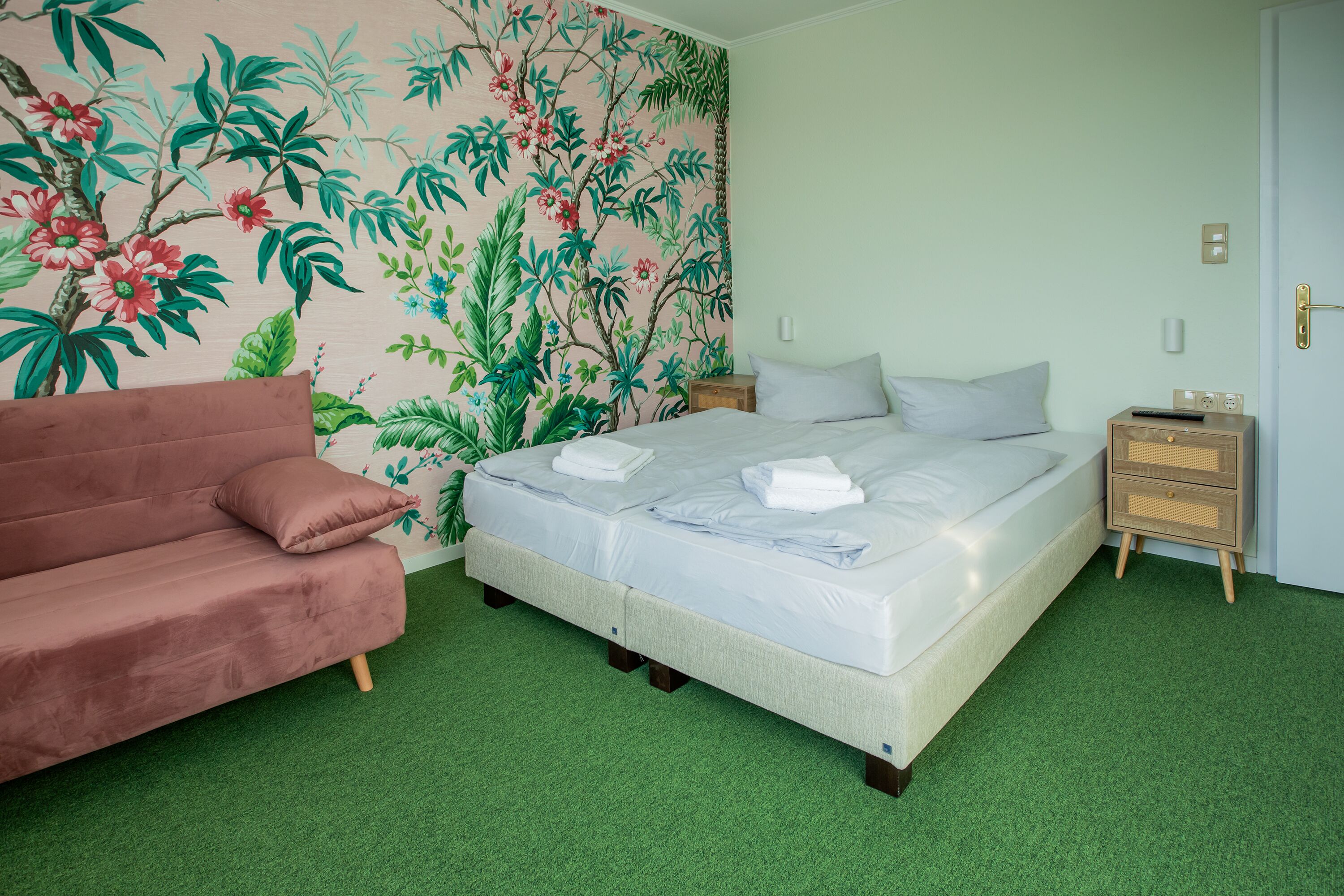 A room with a bed, a couch, a nightstand, and a wall with a floral mural.