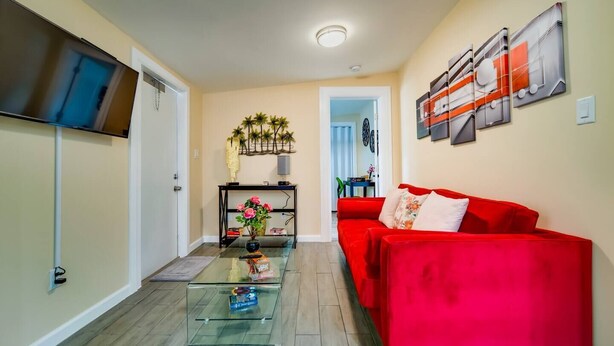 A living room with a red sofa, a glass coffee table, and a TV mounted on the wall. - Small 2BR / Fully Equipped Kitchen / Parking (Fort Lauderdale)
