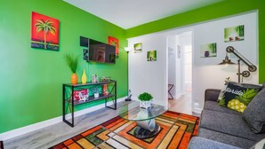 Ein Wohnzimmer mit grüner Wand, einem Glastisch, einem grauen Sofa und einem farbenfrohen Teppich.