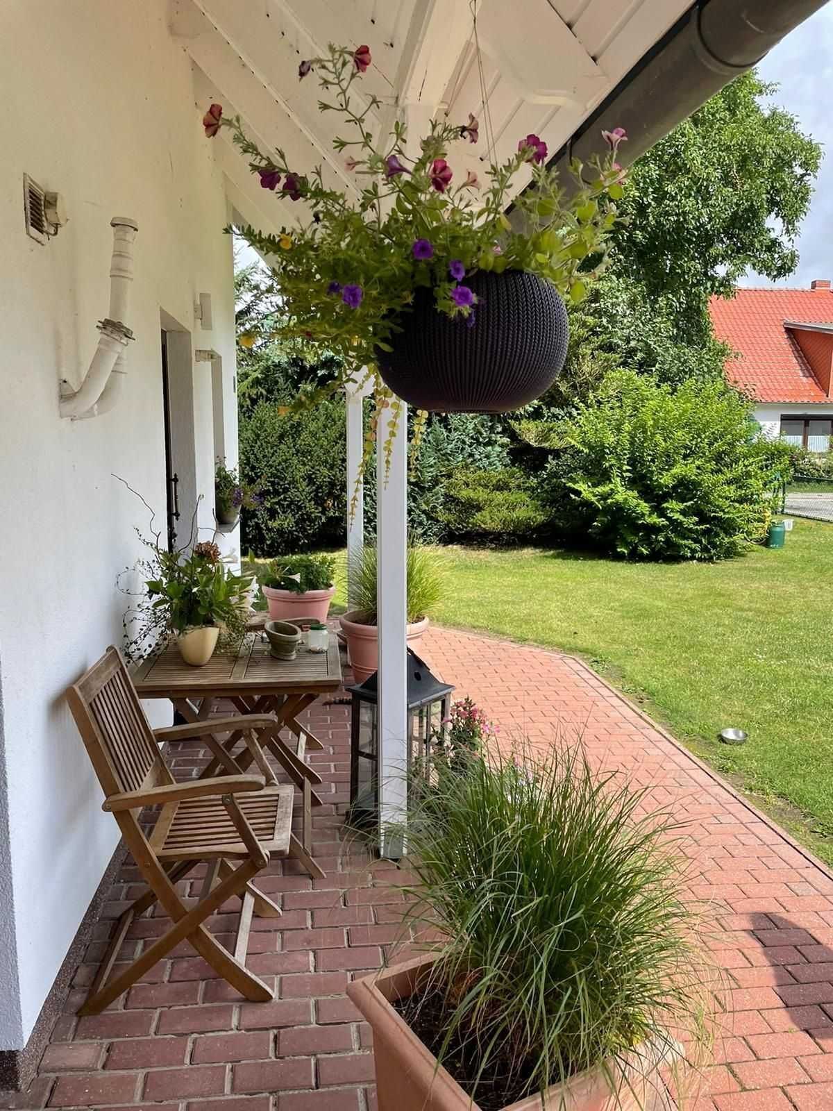 A covered patio with a table and chairs, surrounded by potted plants and a hanging flower basket.
