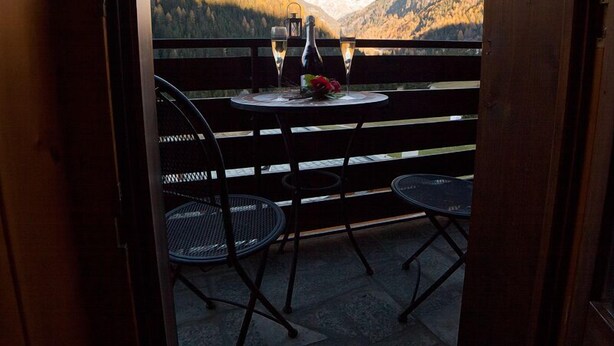 Een balkon met een tafel voor twee, inclusief een fles en glazen, met uitzicht op een bergachtig landschap.