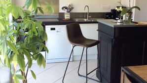 A kitchen with a white dishwasher, a black countertop, a bar stool, a potted plant, and a painting on the wall.