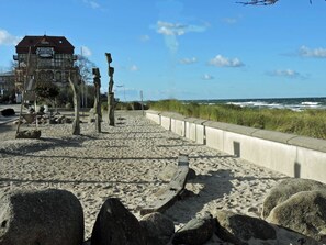 Een strand met een betonnen pad, grote rotsen en een gebouw met een bord 'SCHOOLS - MEER' op de achtergrond.