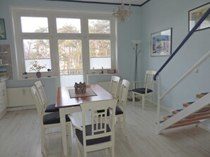Ruokailutila, jossa on puinen pöytä ja valkoiset tuolit, kattokruunu, portaikko ja ikkuna, jossa on sälekaihtimet.