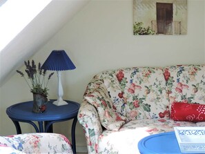 Um quarto com sofá floral, uma mesinha com abajur, planta em vaso e um quadro na parede.