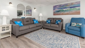 Een woonkamer met twee grijze banken, een blauwe fauteuil, een tapijt met dessin en wanddecoratie van een strandgezicht.
