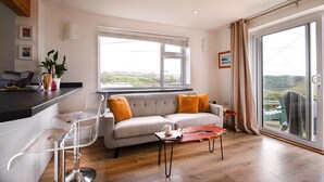 A modern living room with a sofa, two bar stools, a coffee table, and a large window offering a view of the outdoors. - Spectacular one bedroom apartment , incredible coastal views, walk to the  beach (Porthtowan)