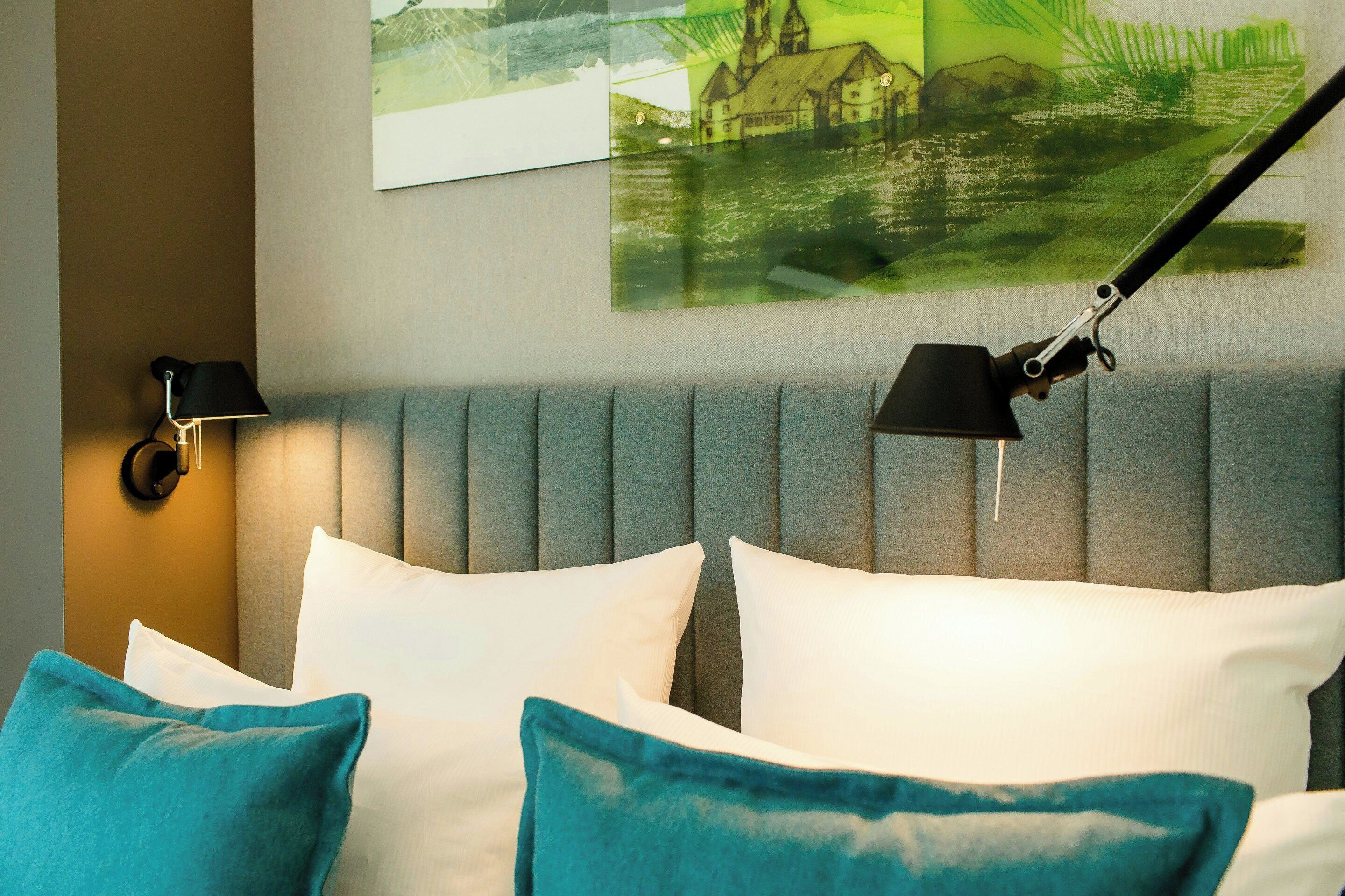 Un lit bien fait, agrémenté d’oreillers blancs et turquoise, d’une tête de lit rembourrée grise et d’une lampe fixée au mur.