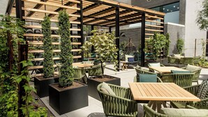 An outdoor patio area with wooden tables and green chairs, surrounded by planters with trees and shrubs. - Motel One Stuttgart-Hauptbahnhof (Stuttgart)