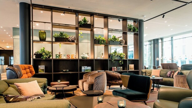 A modern lounge area with comfortable seating, a large bookshelf with plants, and a view of the city.