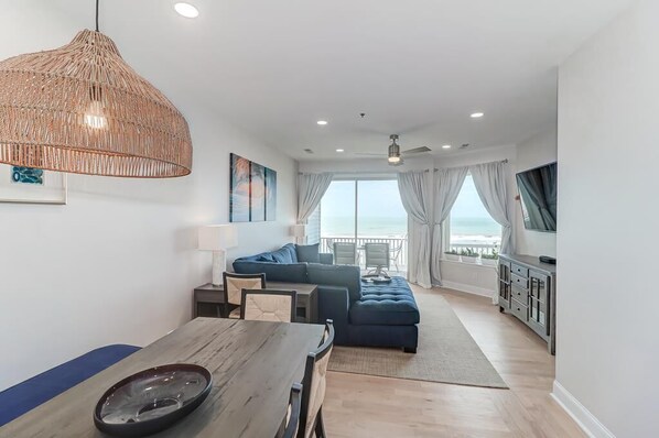Un moderno salón con un ventanal grande con vistas al mar, una zona de comedor con mesa de madera y una acogedora disposición de asientos.
