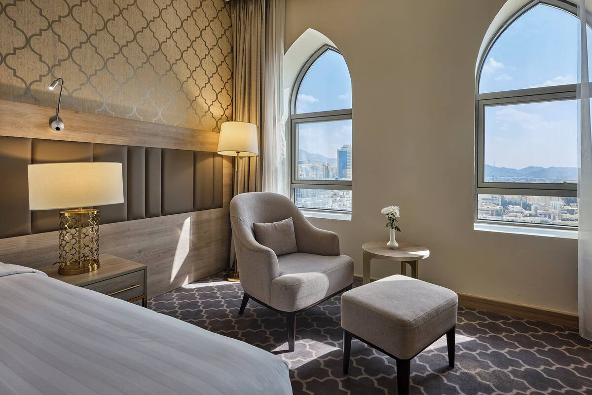 A hotel room with a bed, a chair, a small table, a lamp, and a view of the city through large windows.