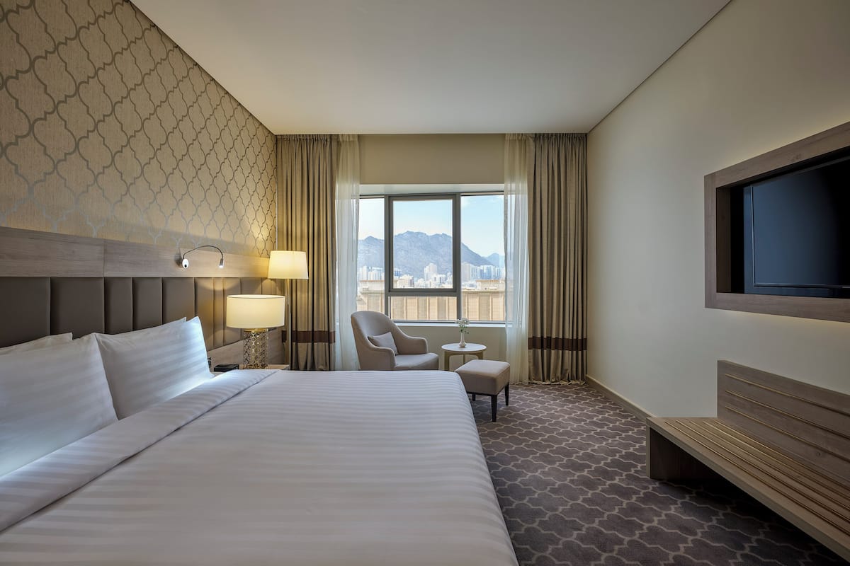 A hotel room with a large bed, a chair, a small table, a TV, and a window with a city view.