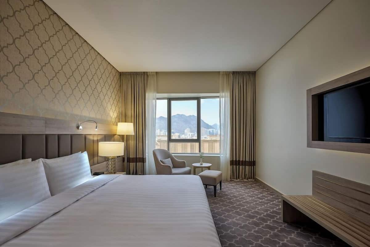 A hotel room with a large bed, a TV, a chair, a small table, and a view of the city.