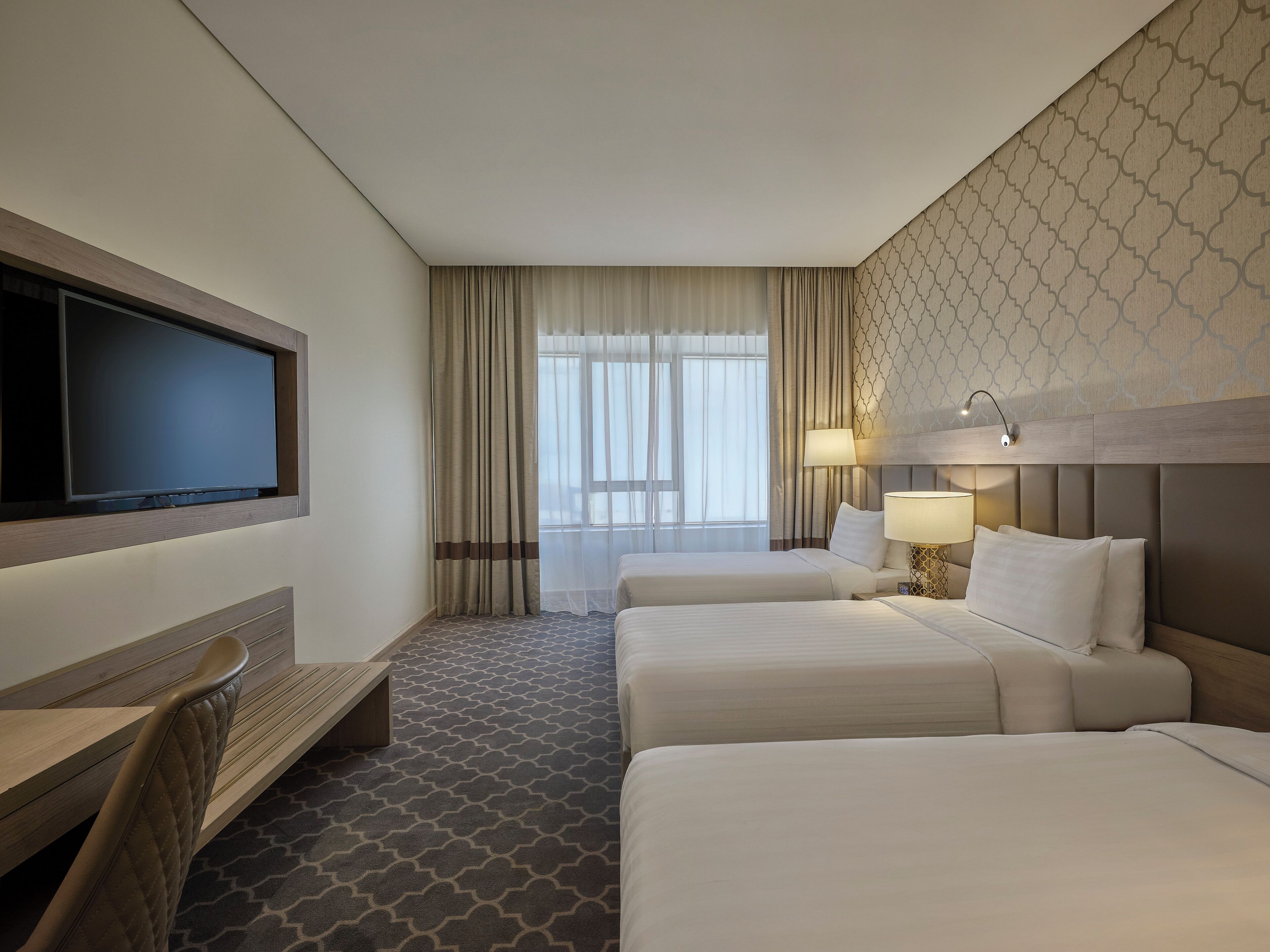 A hotel room with a large flat-screen TV mounted on the wall, two beds with white linens, a wooden bench, a patterned carpet, and a window with sheer curtains.