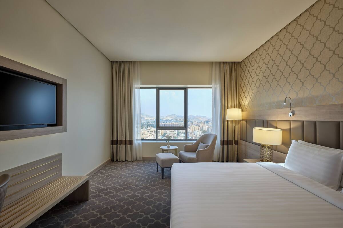 A hotel room with a large bed, a TV mounted on the wall, a desk with a chair, and a view of the city through the window.