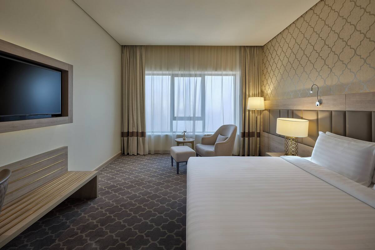 A hotel room with a large bed, a TV mounted on the wall, a desk with a chair, a small table, and a lamp.