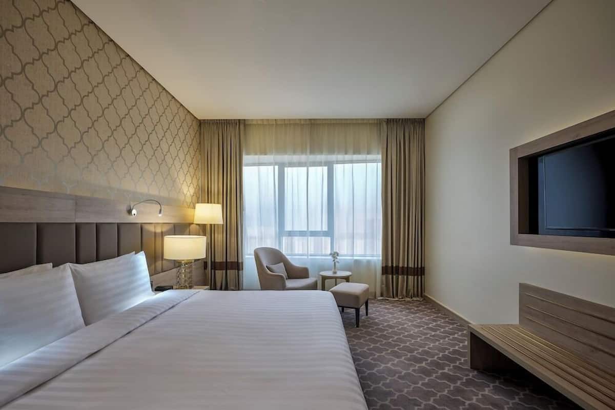 A hotel room with a large bed, a chair, a small table, a TV, and a window with curtains.