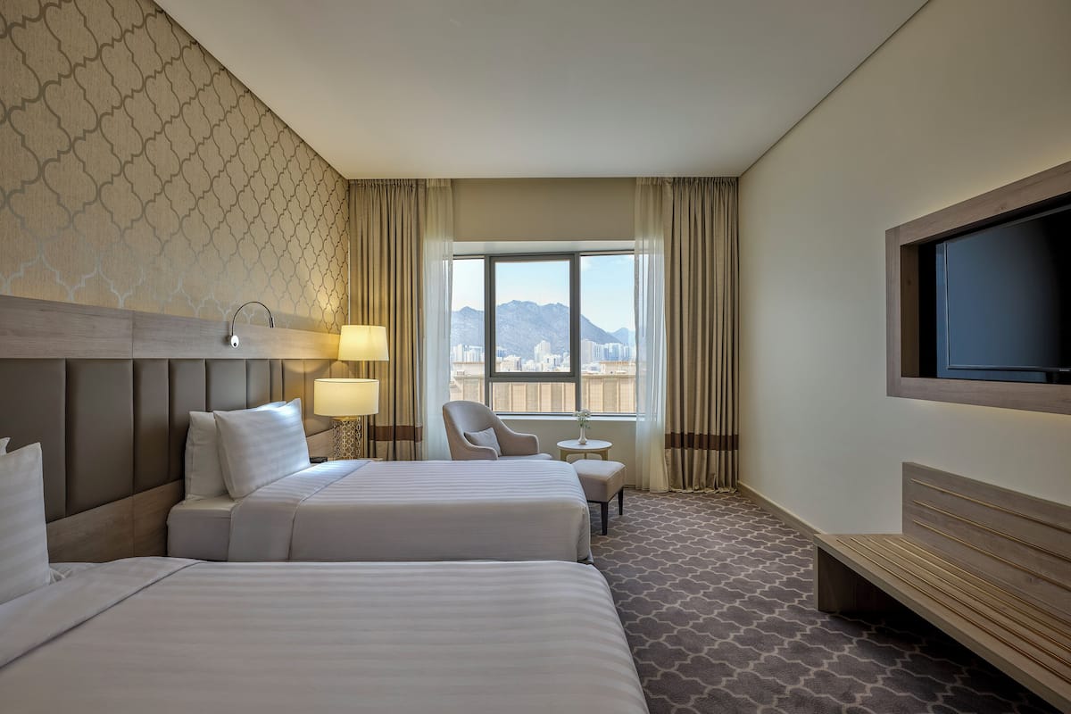 A hotel room with two beds, a TV, a chair, and a window with a city view.