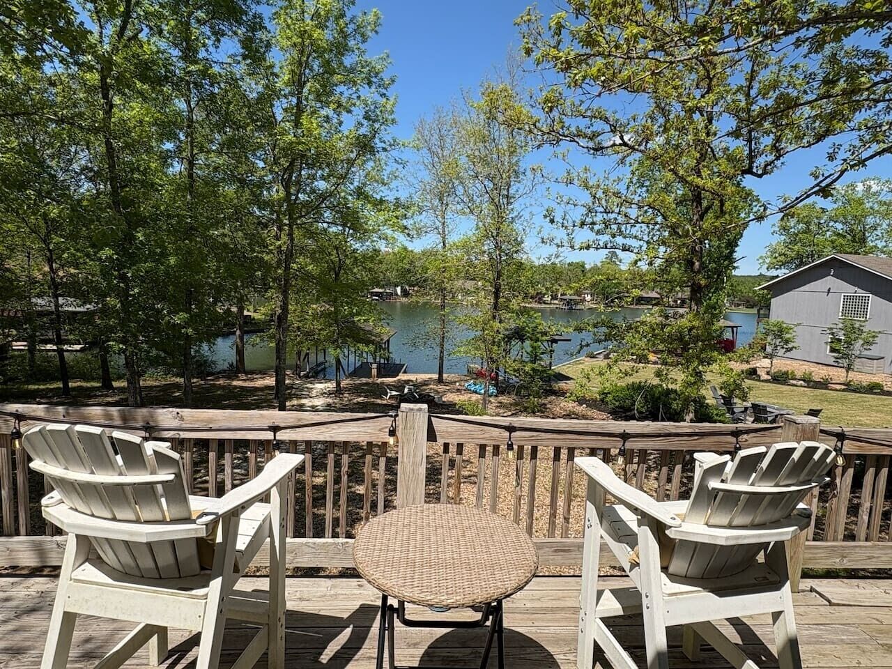 Um terraço com duas cadeiras Adirondack e uma mesa, com vista para um lago com um cais e um edifício ao fundo.