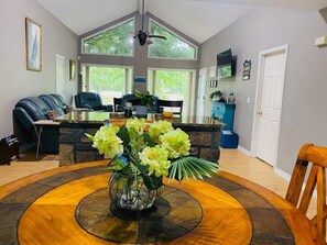 A dining area with a round table, a vase with flowers, a ceiling fan, and a view of the outdoors through large windows.
