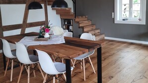 Ein Essbereich mit einem Holztisch, weißen Stühlen und einer schwarzen Pendelleuchte. Im Hintergrund ist eine Treppe mit Holzstufen und einem schwarzen Geländer zu sehen.