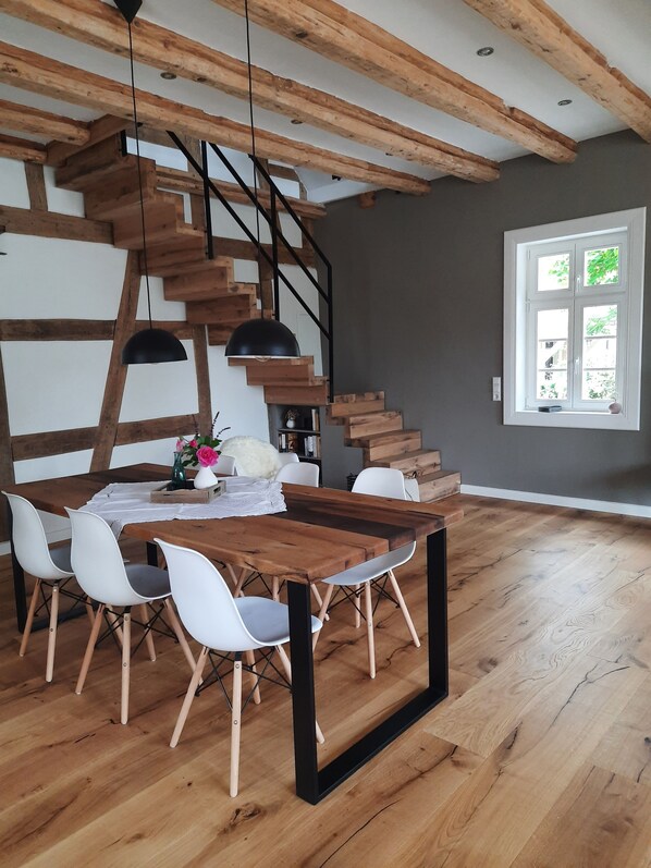 Un coin repas avec une table en bois, des chaises blanches et un luminaire suspendu noir. Un escalier avec des marches en bois et une rampe noire est visible à l’arrière-plan.