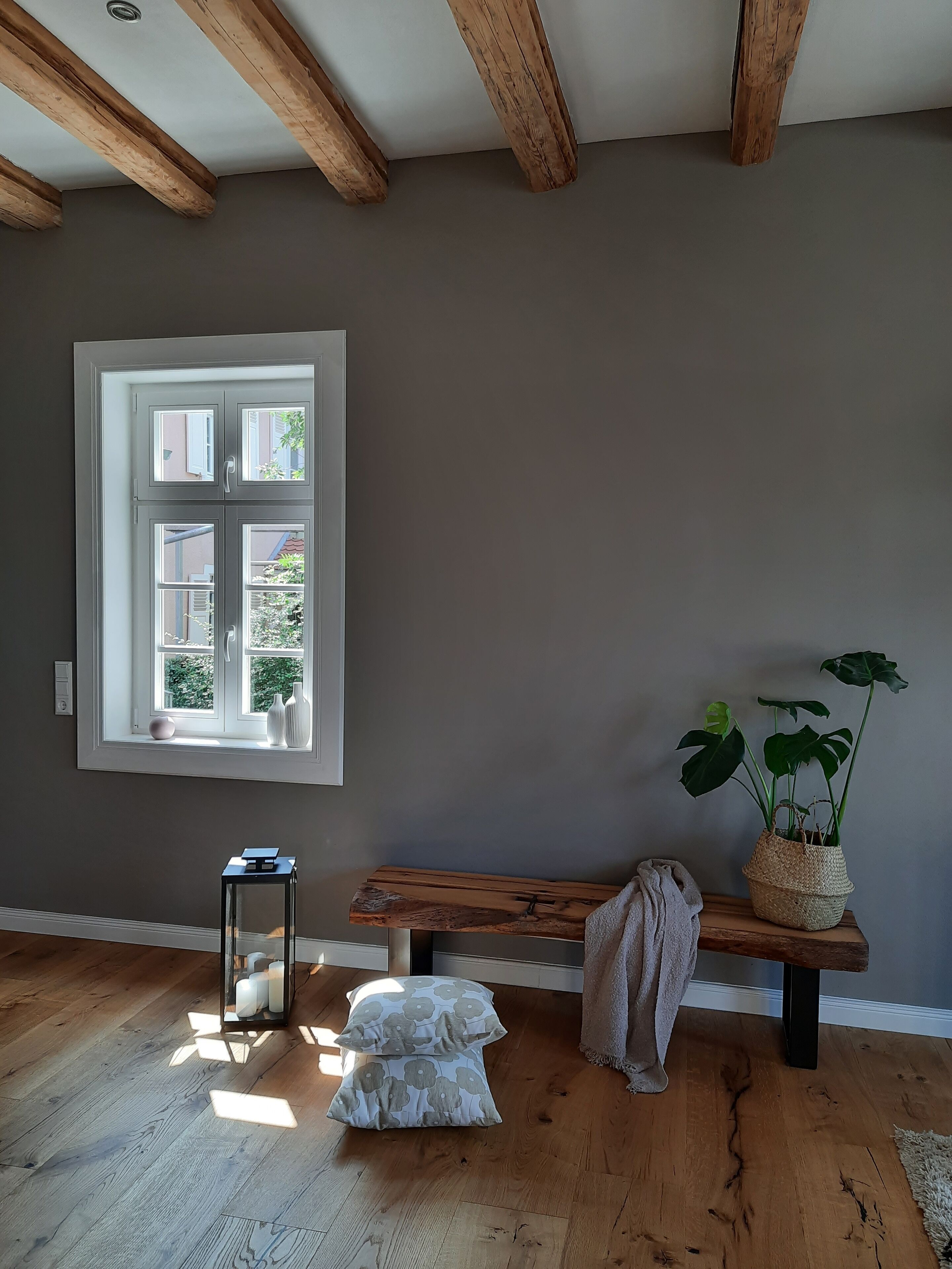 A room with a large window, wooden beams on the ceiling, a bench, a small table, and a potted plant.