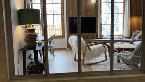 Ein Zimmer mit großem Fenster, einer Holzwand, einem weißen Sofa, einem weißen Sessel, einem kleinen Tisch mit Lampe sowie einem Fernseher.