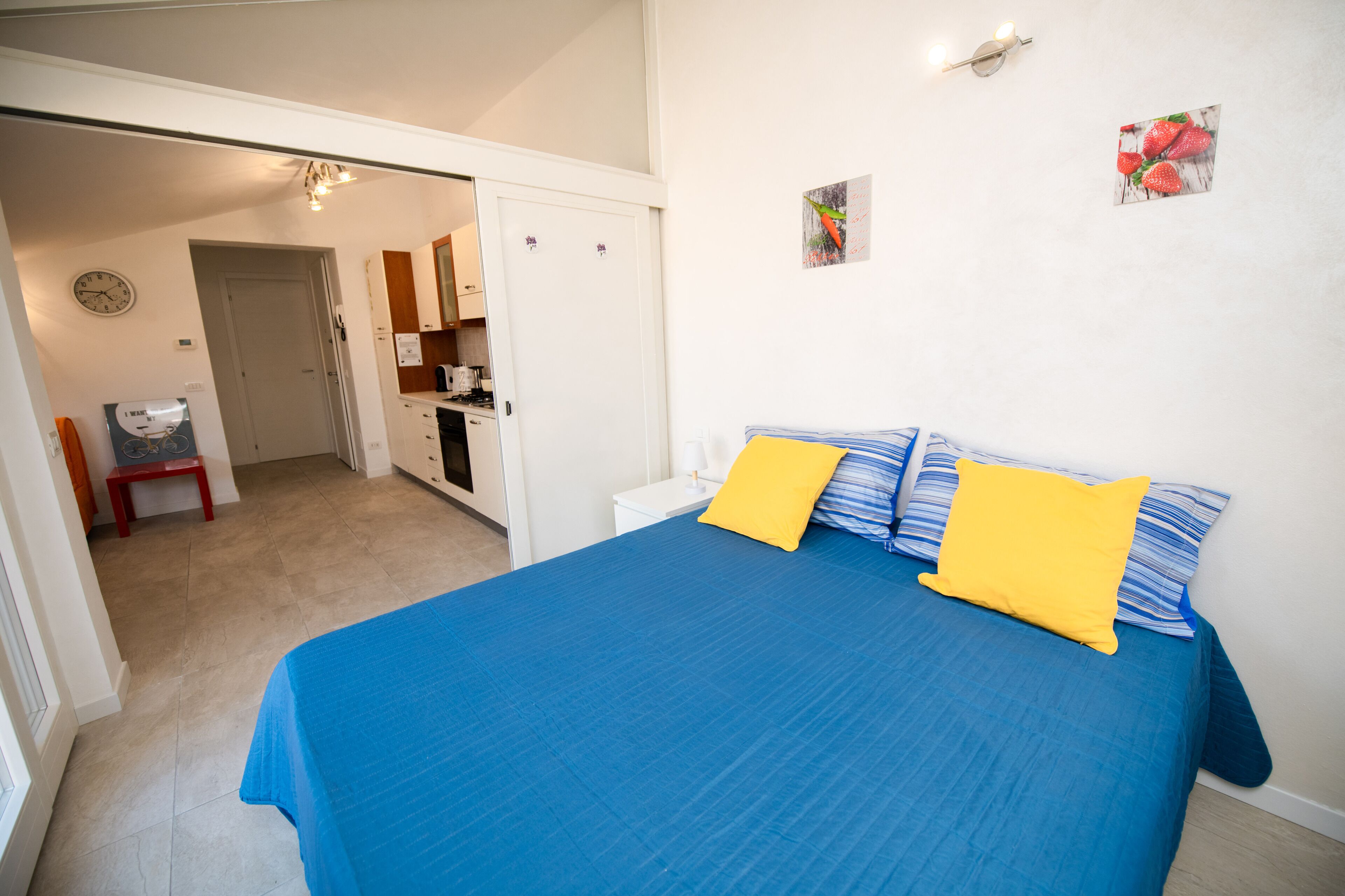 A bedroom with a bed, two pillows, a nightstand, a wall clock, a small kitchen area, and a door leading to another room.