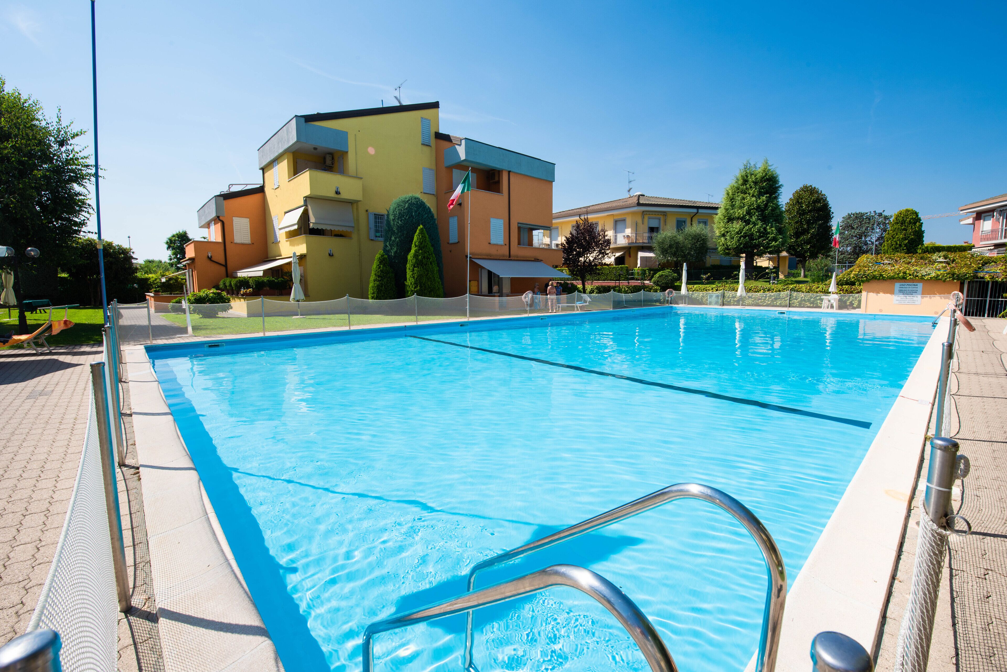 Ein Schwimmbecken mit einer Metallleiter, umzäunt und neben einem mehrstöckigen Gebäude mit Balkonen.