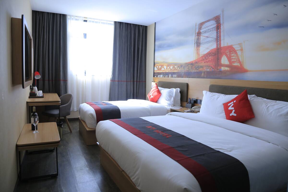 A hotel room with two beds, a desk with a chair, a small table with a wine glass, and a painting of a bridge.