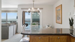 Une cuisine moderne dotée d’un îlot central, d’un coin repas avec une table en verre et offrant une vue sur l’océan grâce à de grandes fenêtres.