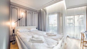 A hotel room with a large bed, a bedside table with a lamp, a view of the city through the window, and a balcony with a chair. - Apartamenty Sun & Snow Riverside Sienna (Gdansk)