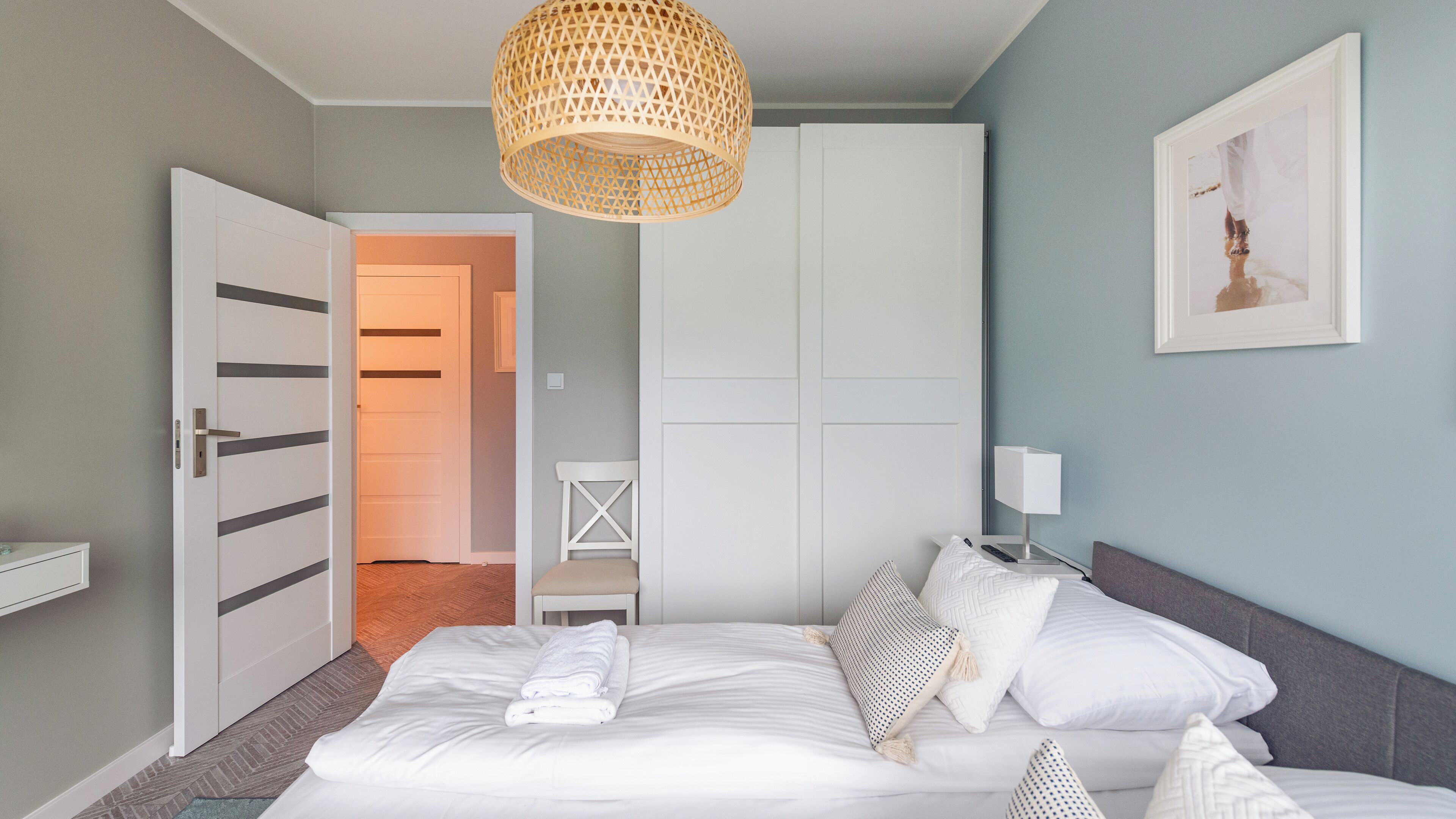 Un dormitorio con cama, paredes blancas, una puerta de madera, un armario blanco, una mesita de noche con lámpara y un cuadro enmarcado en la pared.