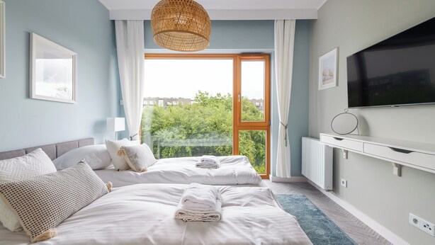 A modern hotel room with two beds, a large window with a view of greenery, a flat-screen TV, and a white dresser. - Apartamenty Sun & Snow Riverside Sienna (Gdansk)