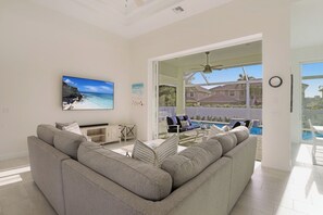 A modern living room with a large gray sofa, a flat-screen TV, and a view of a pool and houses through an open door. - Beachy Home | Pool and Steps to Beach/Food/Shops (Marco Island)