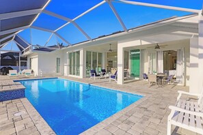 A swimming pool with clear blue water, surrounded by a covered patio area with white furniture and a glass-enclosed room. - Beachy Home | Pool and Steps to Beach/Food/Shops (Marco Island)