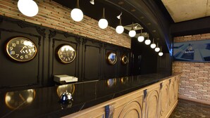 A reception area with a black counter, multiple wall clocks, hanging pendant lights, and a brick wall backdrop. - Bayberd Hotel (Bayburt)