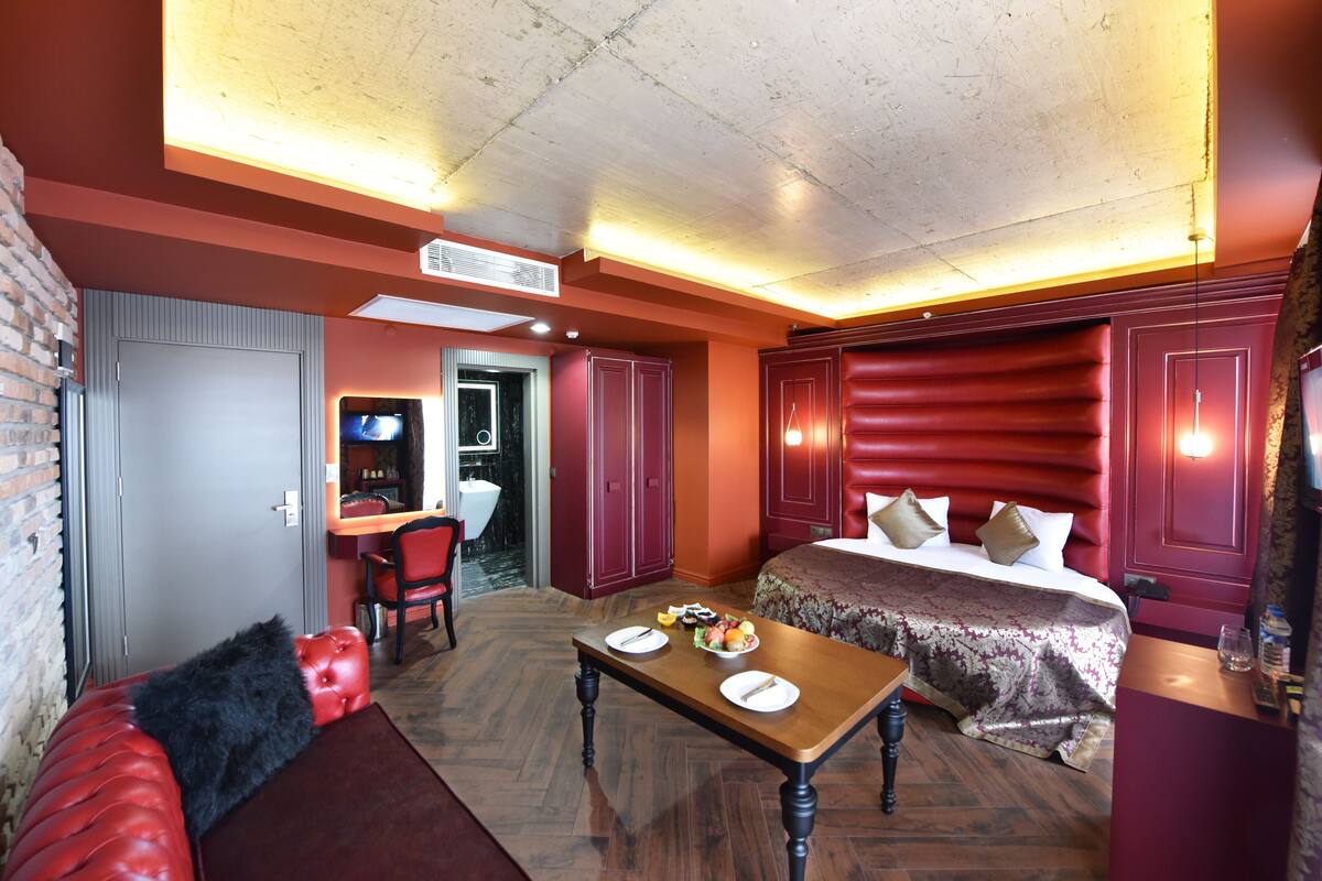 Una habitación de hotel con cama, una mesa de comedor con platos y cubiertos, un sofá de cuero rojo y una pared de ladrillo.