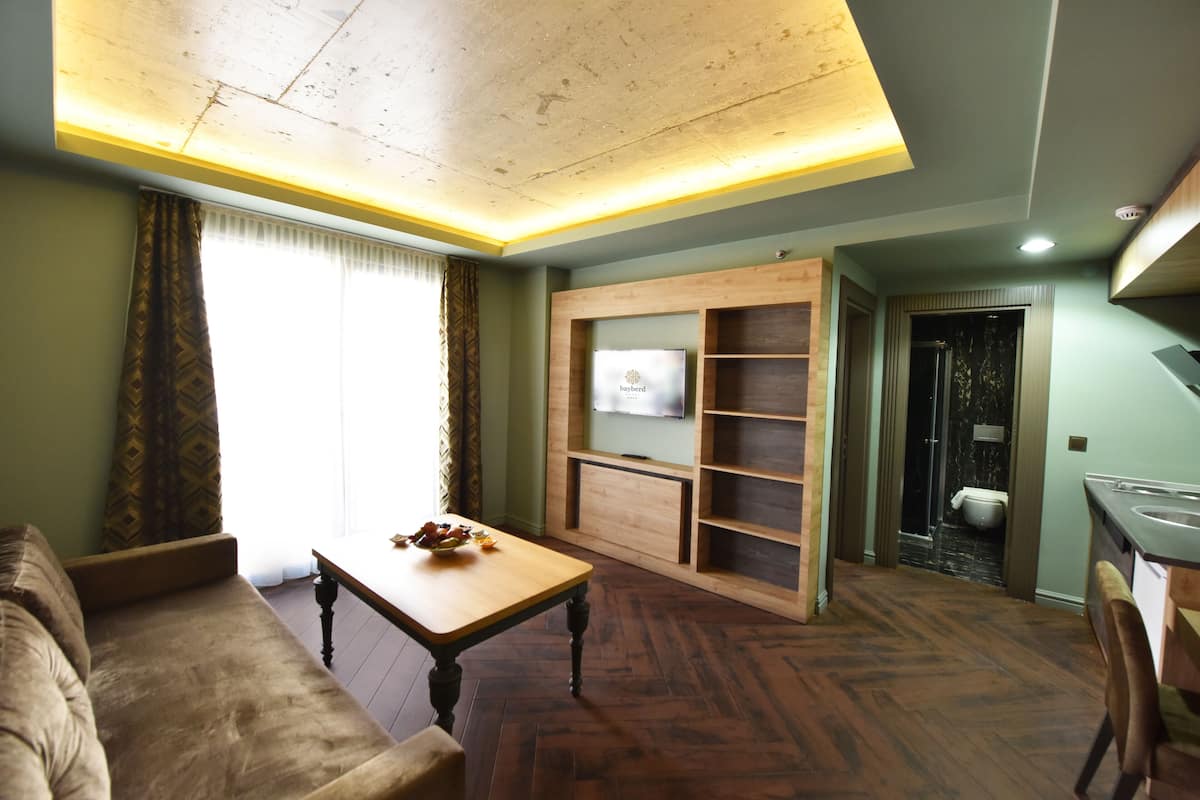 Una sala de estar con una mesa de centro de madera, un sofá, un televisor montado en una pared de madera, y un baño visible a través de una puerta abierta.
