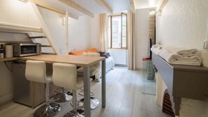 Une chambre d’hôtel compacte comprenant un coin cuisine, un espace repas avec deux tabourets de bar, un lit avec une couette blanche et une fenêtre avec des rideaux.